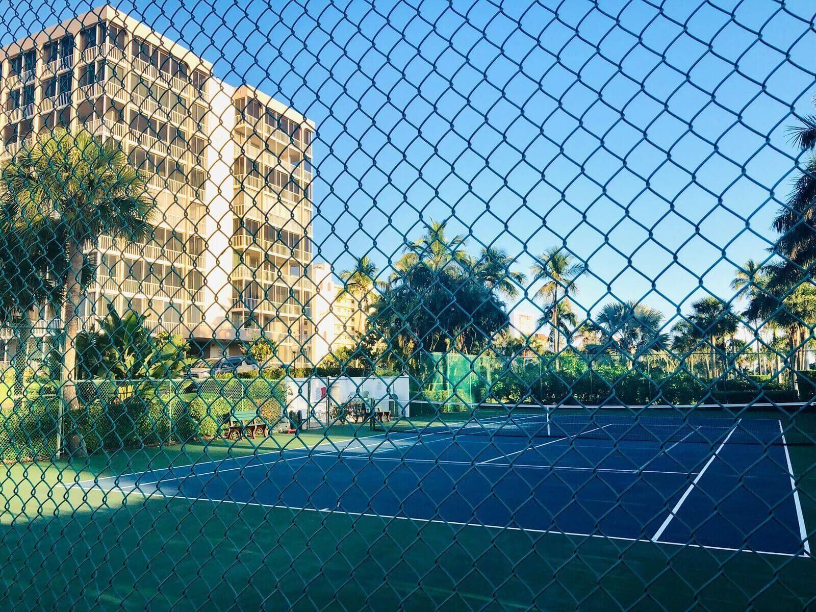 A chain-link fence surrounding a tennis court with a building in the background.