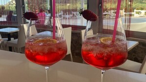 Dos vasos de bebida roja con pajitas y adornos sobre una mesa en un restaurante con ventanas grandes y cortinas rojas.
