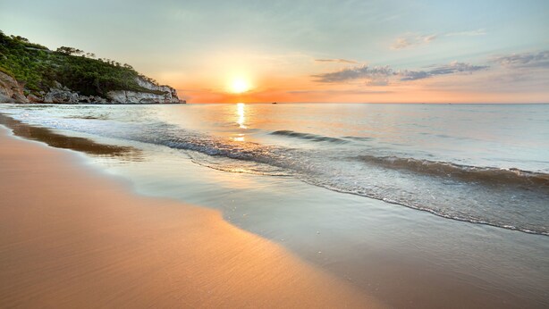 Una playa con un atardecer, aguas tranquilas y un acantilado rocoso.