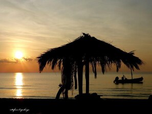 A beach scene with a thatched hut and a boat in the water during sunset.