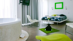 Una habitación de hotel moderna con una bañera grande, un área de estar con un otoman verde, una cama con almohada verde y un televisor de pantalla plana montado en la pared.