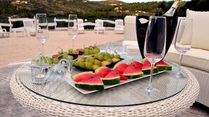 Una mesa con cubierta de vidrio, rodeada de sillas de mimbre, con un plato de sandía, uvas y una botella de champán.