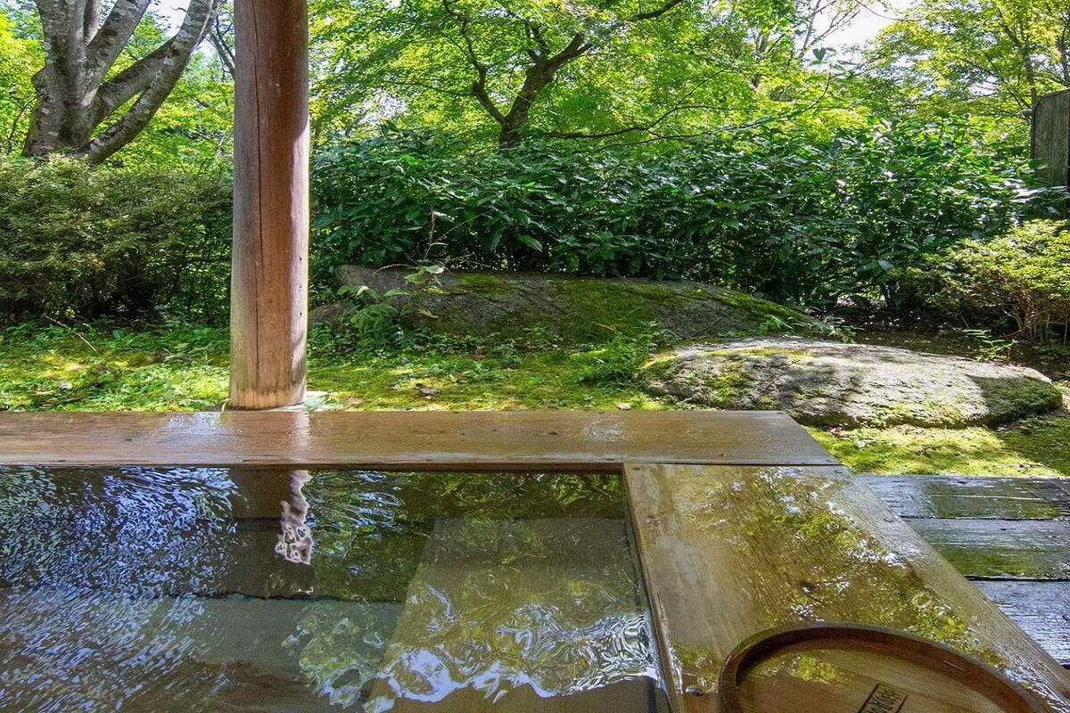 Un balneario de aguas termales de madera con borde de piedra, rodeado de vegetación exuberante y una valla de madera.