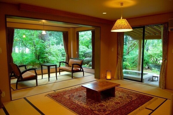 Una habitación con suelo de tatami, una mesa de madera baja, dos sillas y un gran ventanal corredero que da a un jardín.