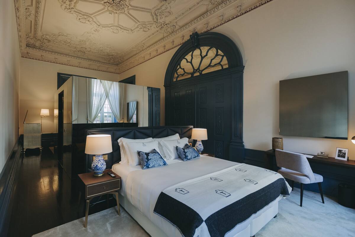 Une chambre d’hôtel avec un grand lit, deux tables de chevet avec des lampes, un bureau avec une chaise, un grand miroir et un téléviseur fixé au mur.