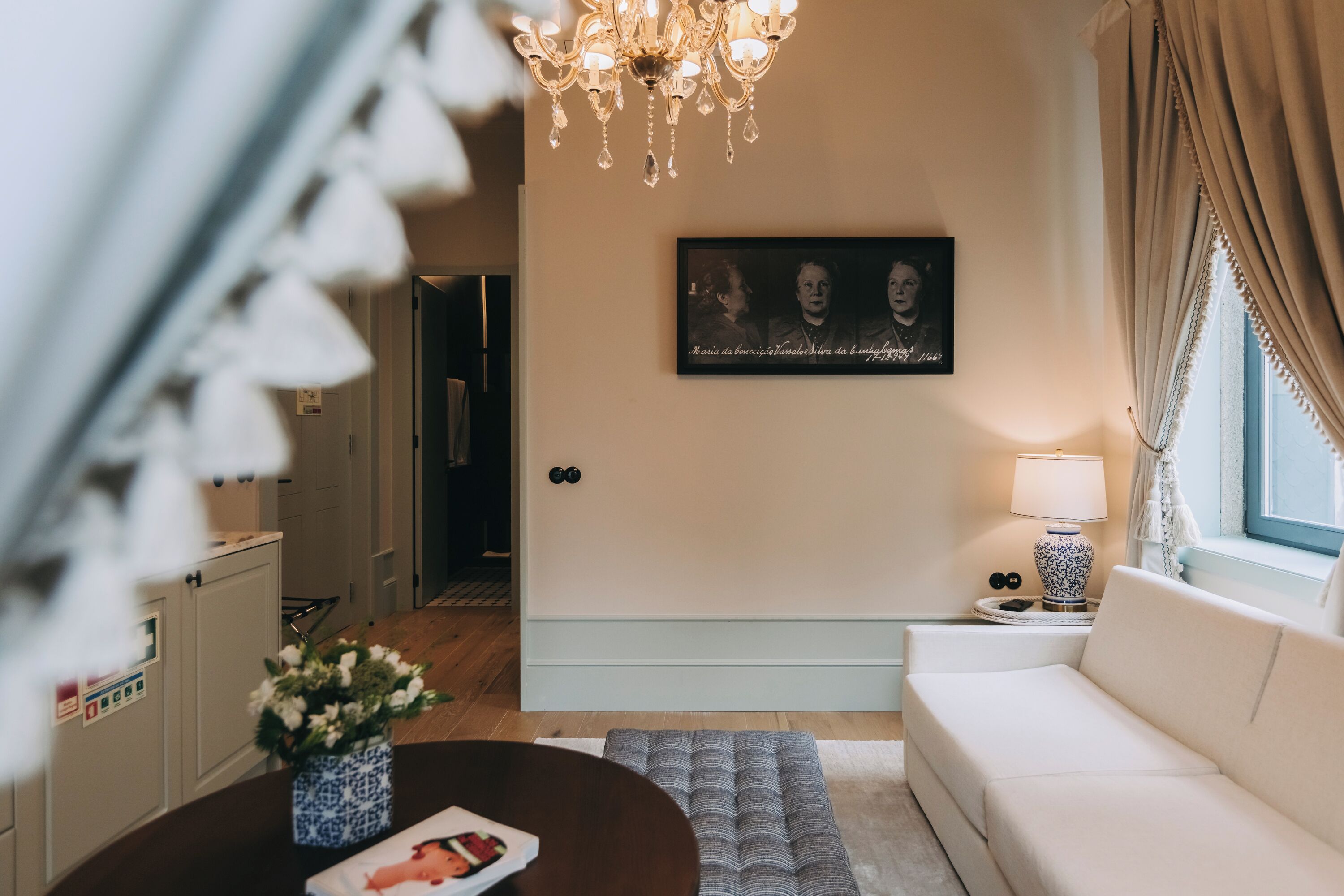 A living room with a white sofa, a round coffee table with a vase of flowers, a chandelier, and a framed picture on the wall.
