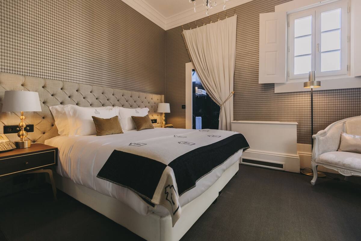 Une chambre d’hôtel avec un grand lit, une table de chevet avec une lampe, une chaise et une vue sur l’extérieur par une fenêtre.