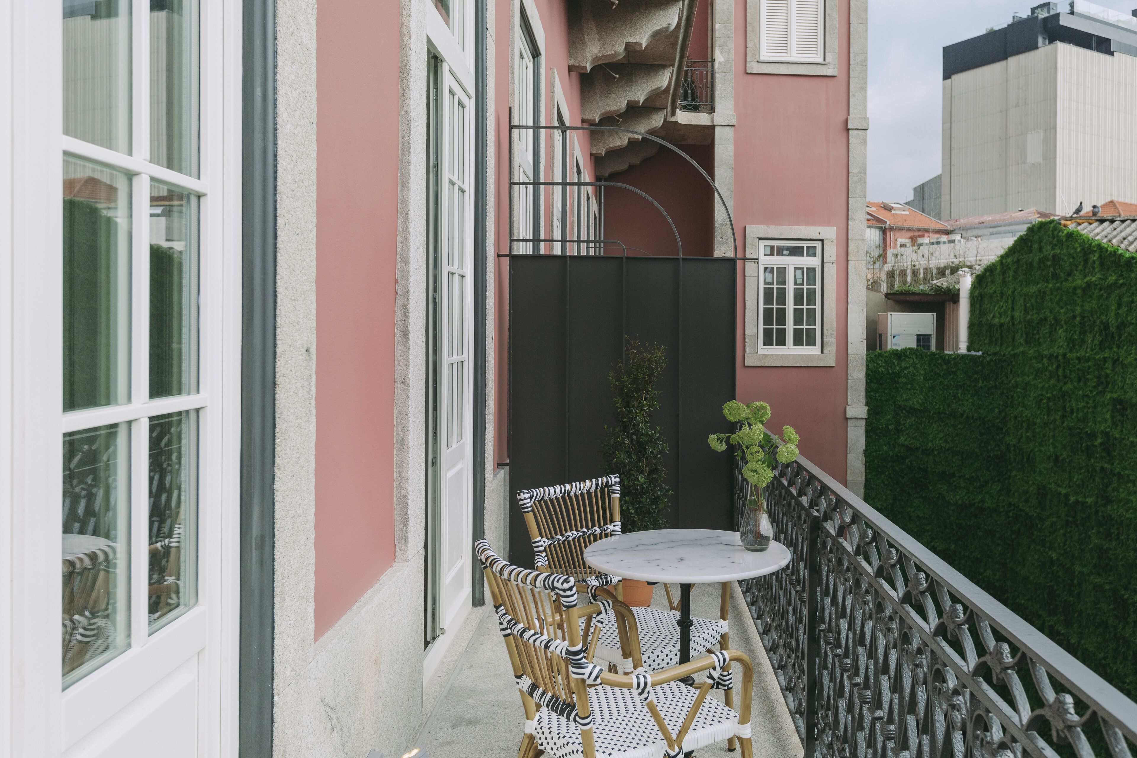 A balcony with wicker chairs and a small table, a potted plant, and a view of a green hedge and a building in the background.