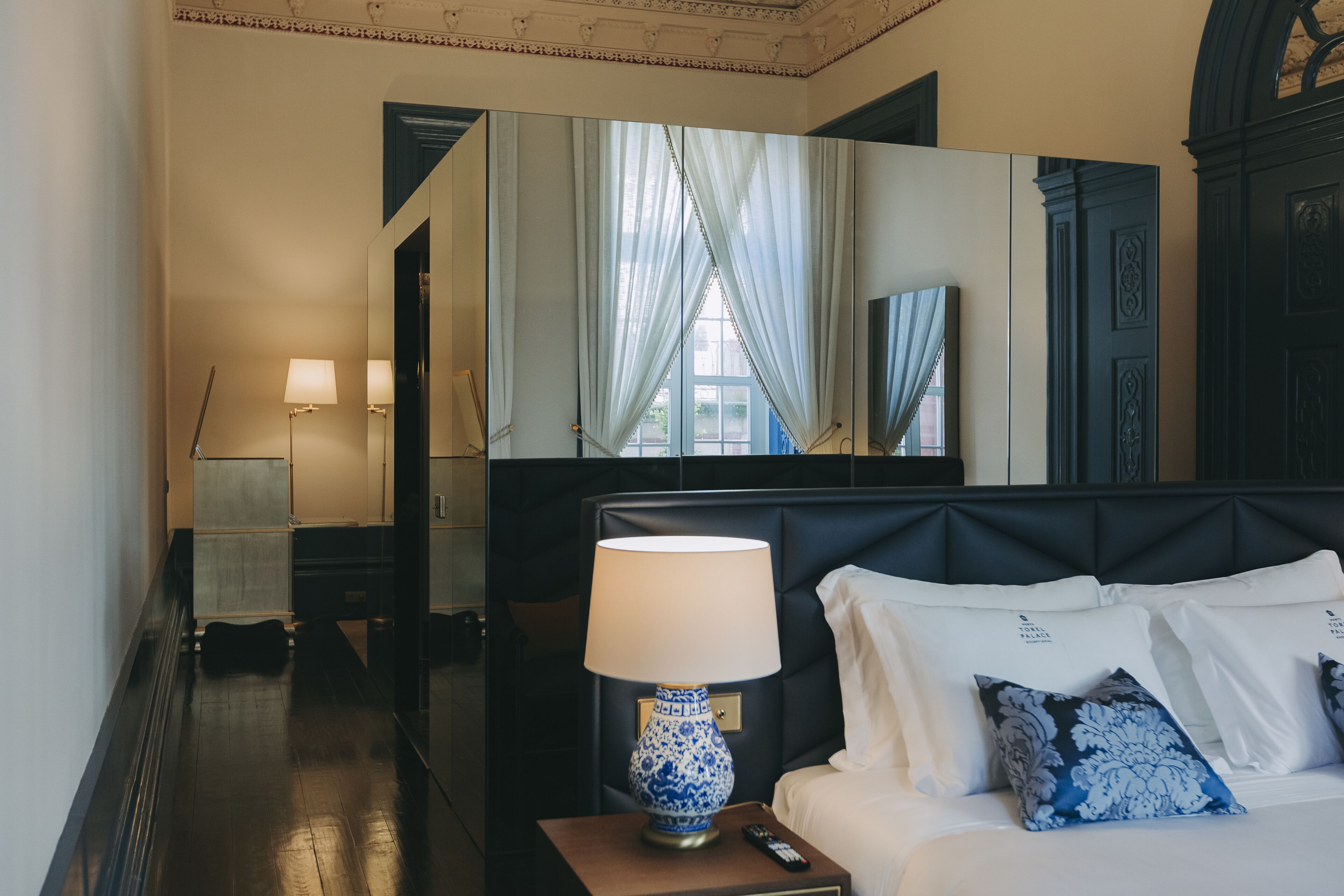 Habitación de hotel con cama, mesita de noche con lámpara, espejo y vista al exterior por una ventana.