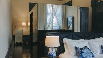 Habitación de hotel con cama, mesita de noche con lámpara, espejo y vista al exterior por una ventana.