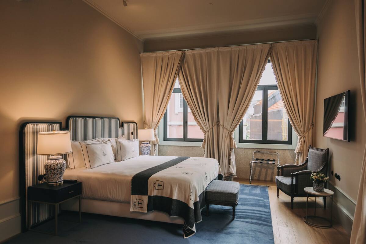 Une chambre d’hôtel avec un grand lit, deux tables de chevet avec des lampes, une chaise, une petite table avec des fleurs et une vue sur les bâtiments par la fenêtre.