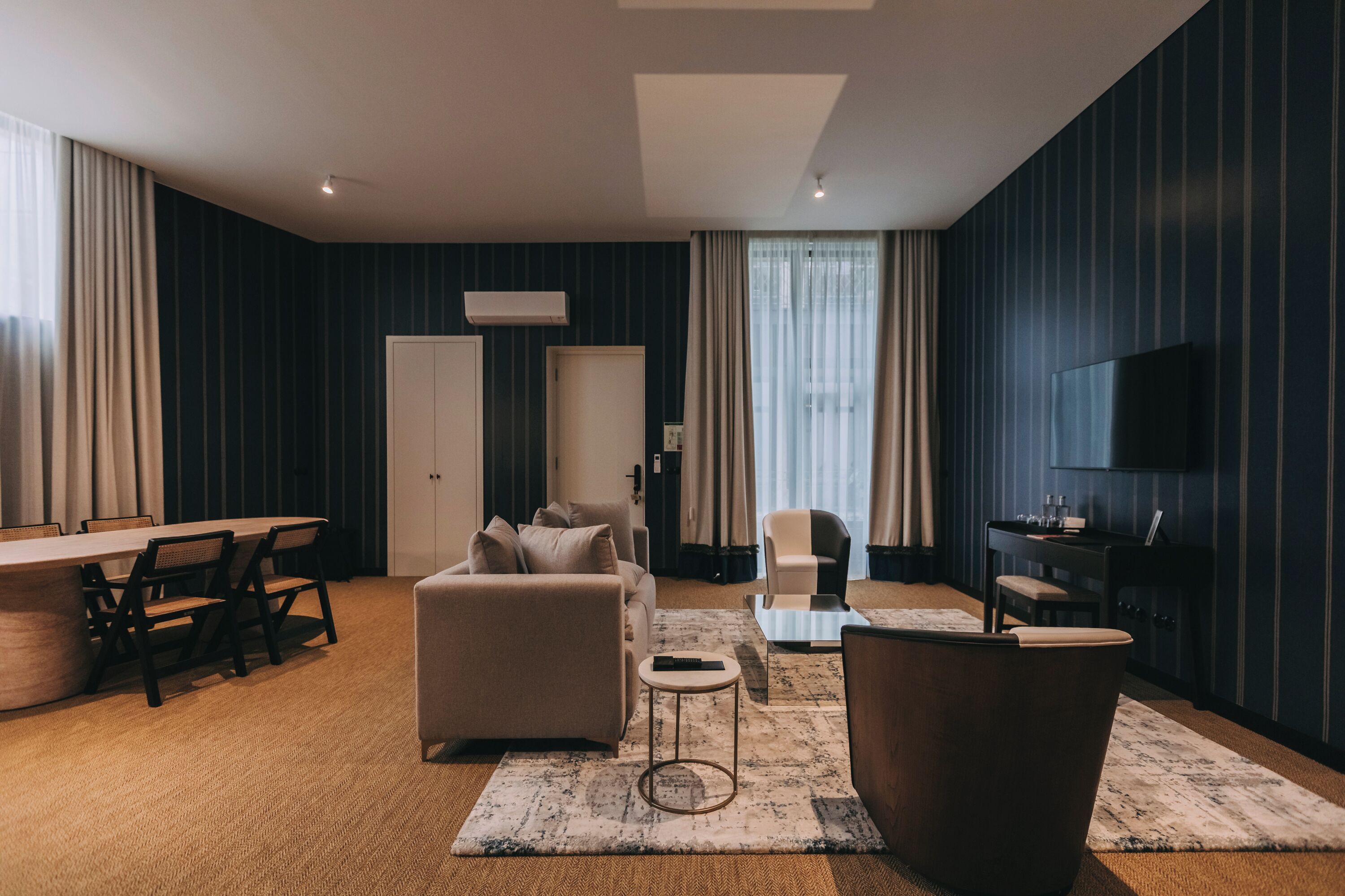 A modern hotel room with a sofa, armchair, dining table, and a TV.
