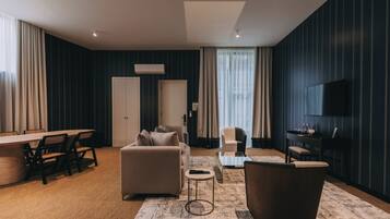 A modern hotel room with a sofa, armchair, dining table, and a TV.