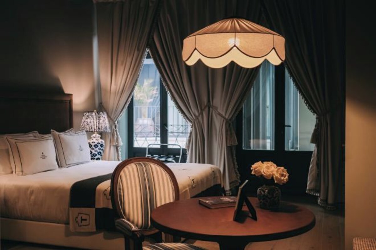 Quarto de hotel com cama, mesa redonda com um vaso de flores, uma cadeira, abajur e cortinas.