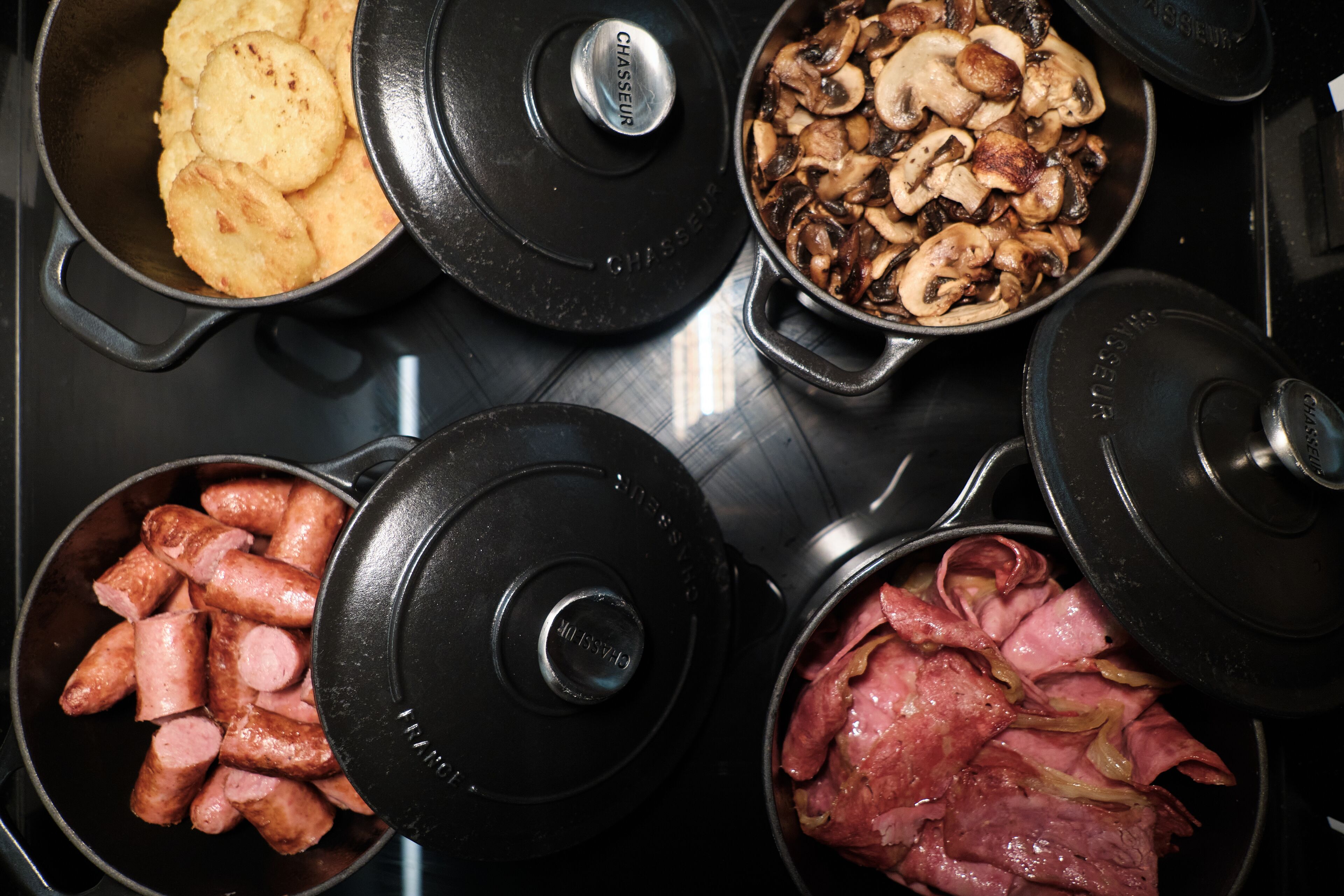Trois casseroles sur une cuisinière, contenant différents aliments : des saucisses, des champignons et du jambon.