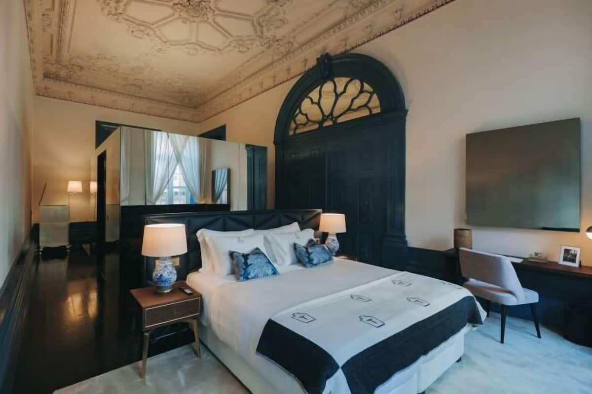 Une chambre avec un grand lit, deux tables de chevet avec des lampes, une chaise, un bureau, un grand miroir et un téléviseur fixé au mur.