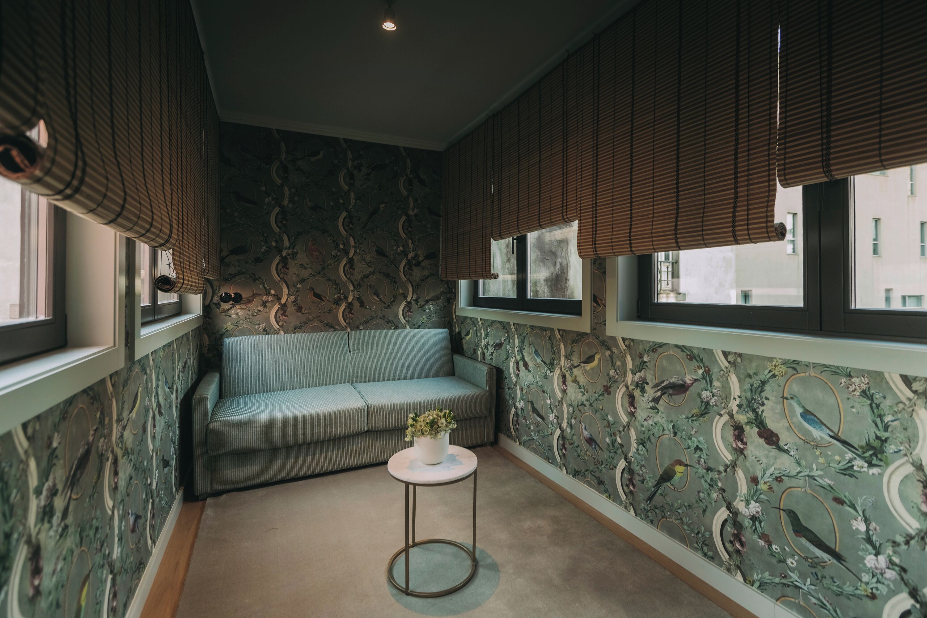 A room with a sofa, a small round table with a plant, patterned wallpaper, and blinds on the windows.