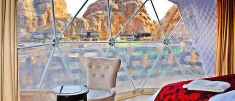 A room with a view of rocky terrain through a transparent dome structure. There is a bed with a red floral bedspread and a white pillow, a small round table with a chair, and a second chair with a light-colored cushion.