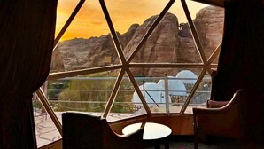 A room with a large window offering a view of rock formations and a sunset. There are two chairs and a small table. - Seven Wonders Luxury Camp (Wadi Musa)