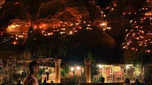 A person sitting on a stone ledge at night, overlooking a dining area with tables and chairs, and a fire pit in the foreground. - Seven Wonders Luxury Camp (Wadi Musa)