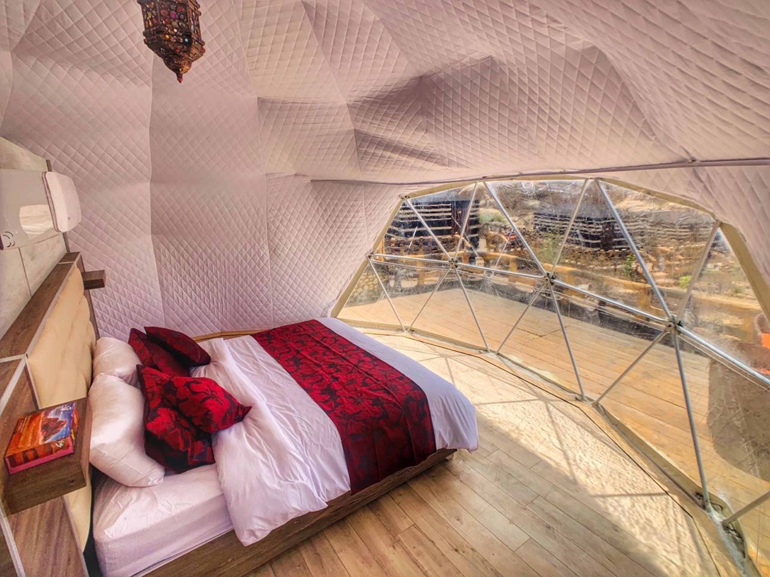 A tented room with a bed, a wooden floor, and a view of a rocky landscape through a glass window.