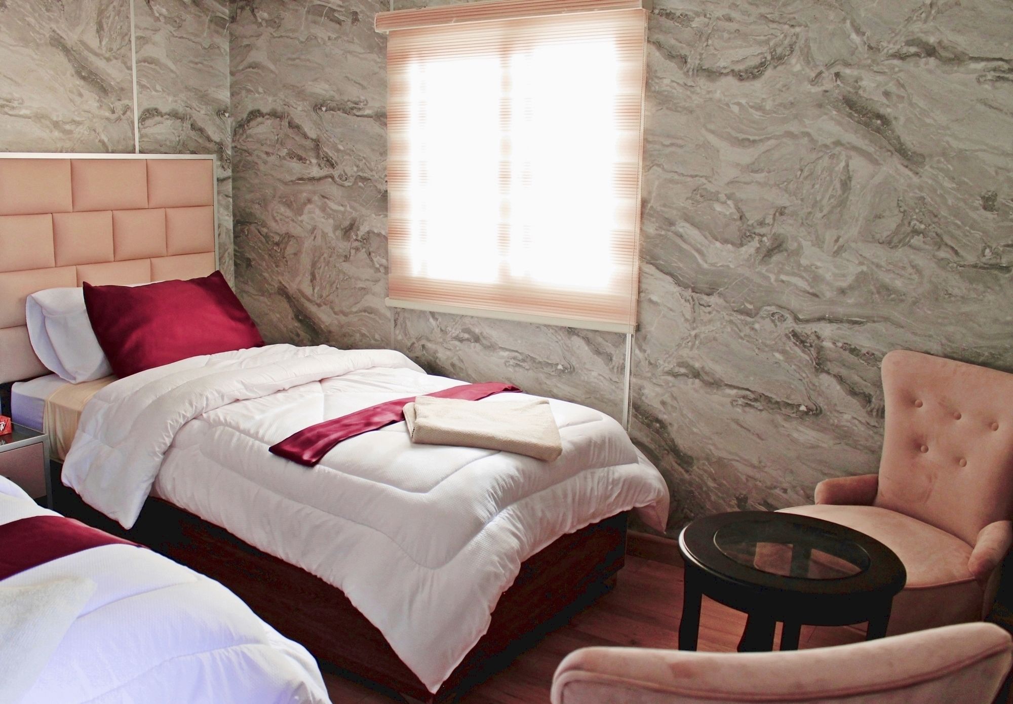 A hotel room with a marble wall, a bed with white bedding and red accents, a chair, and a small table.