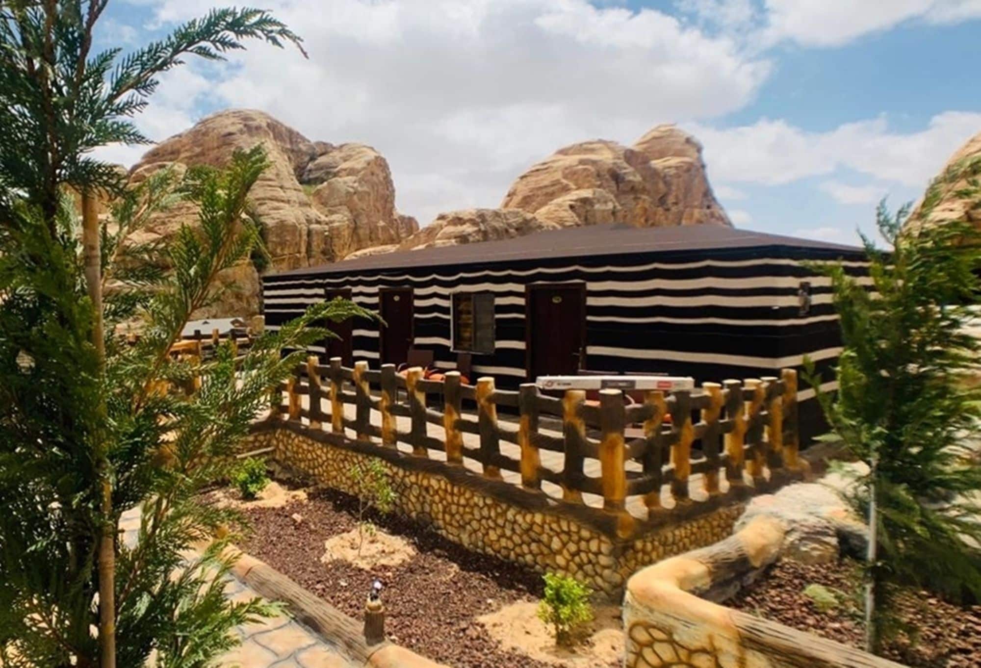 A building with a striped exterior, surrounded by a stone wall and a wooden fence, set against a backdrop of rocky hills.
