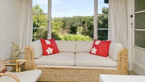 A room with a wicker sofa, two red pillows with white floral patterns, a small wicker chair, and a white table.