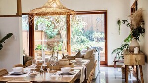 Un coin repas avec une table en bois dressée pour quatre personnes, entourée de chaises en osier. Un luminaire suspendu en fibres tressées se trouve au-dessus de la table, et une petite table d'appoint en bois, ornée d'un vase et d'une bougie, est placée à droite.
