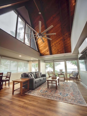 A spacious living area with a high ceiling, large windows, a ceiling fan, and a view of the outdoors.