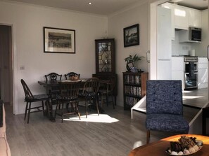 Un espace repas comprenant une table et des chaises en bois, une bibliothèque et un coin cuisine équipé d’appareils électroménagers.