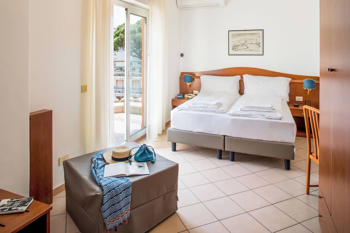 A hotel room with a bed, a desk, a chair, a window with a view of the outside, and a wall-mounted lamp.