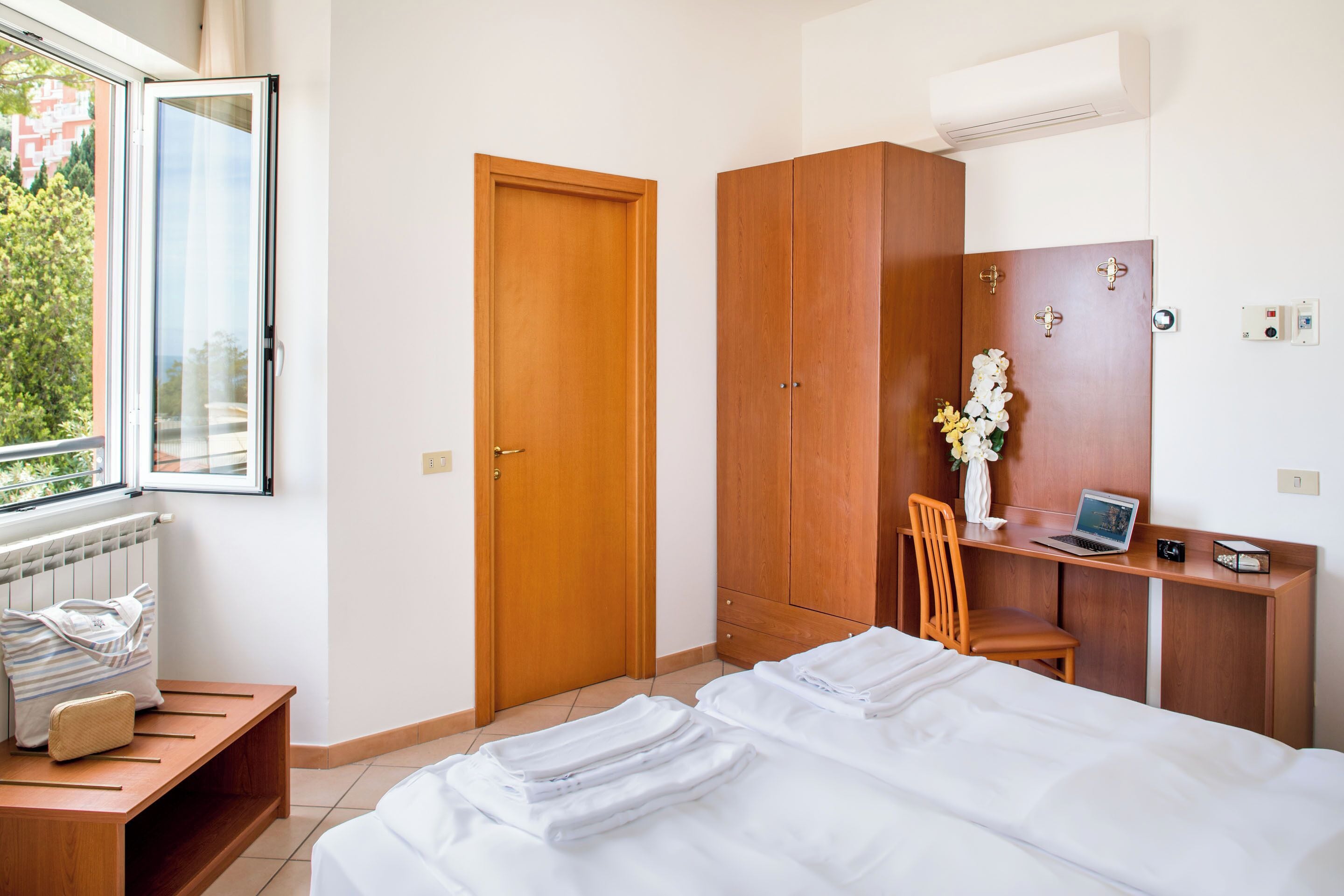 Habitación de hotel con cama, escritorio con una computadora portátil, silla, armario de madera y ventana con vista a follaje.