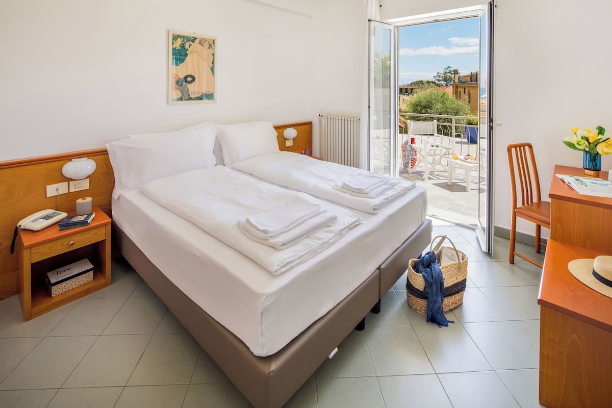 A hotel room with a bed, a bedside table with a lamp, a chair, a small table with a vase of flowers, and a view of the outside balcony.