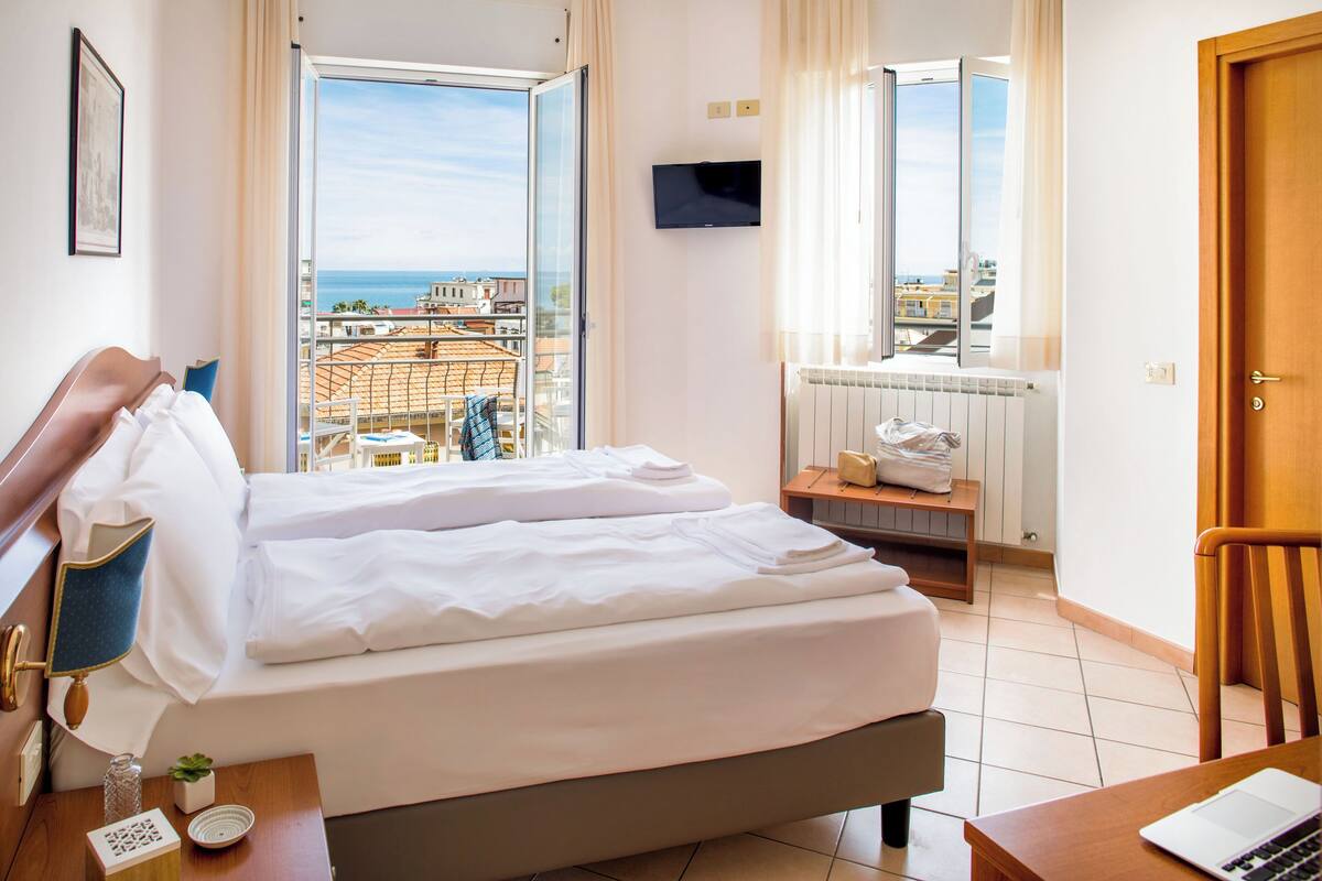 A hotel room with a bed, a desk with a laptop, a TV, and a view of the city and sea.