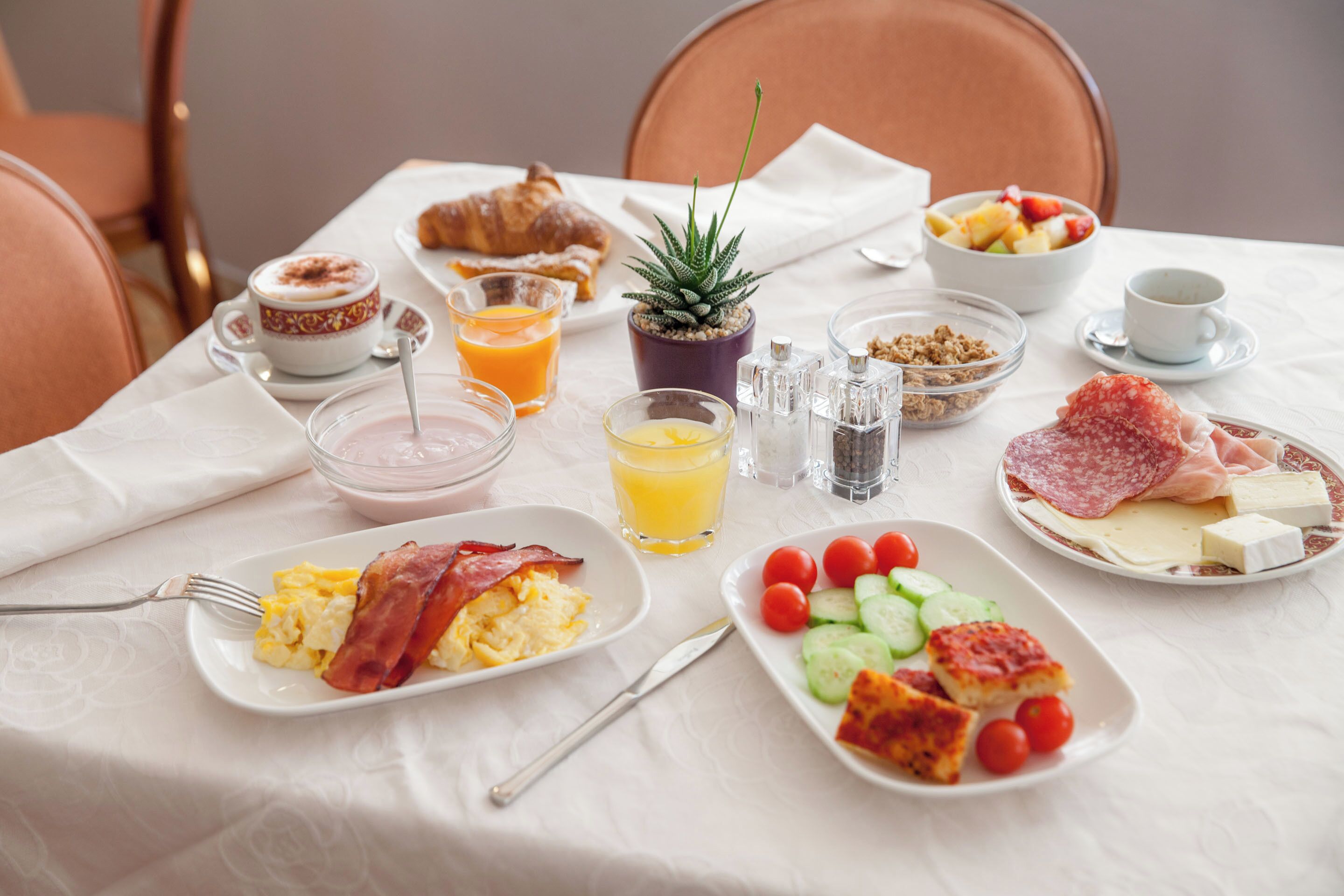 A breakfast table with croissants, eggs, bacon, tomatoes, cucumbers, cheese, and a small plant.