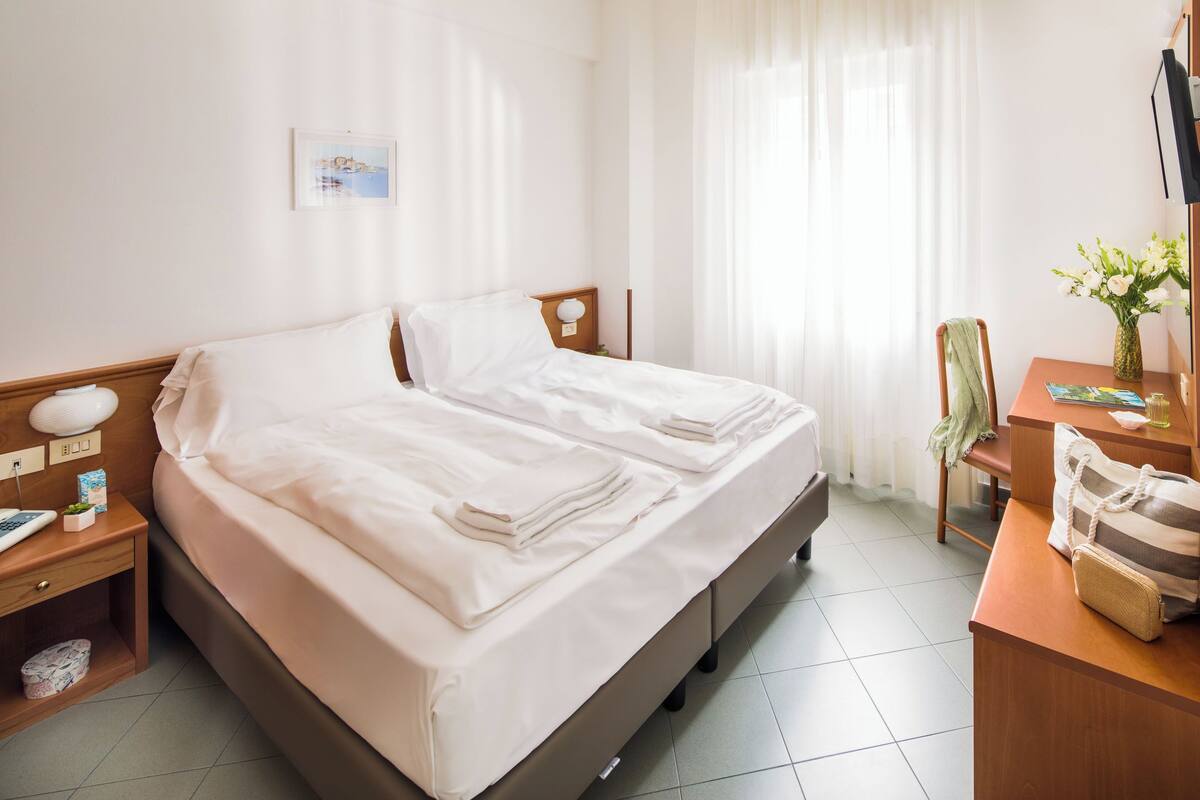 A neatly made double bed with white linens, a wooden nightstand with a lamp, a small table with a vase of flowers, and a chair with a green towel.