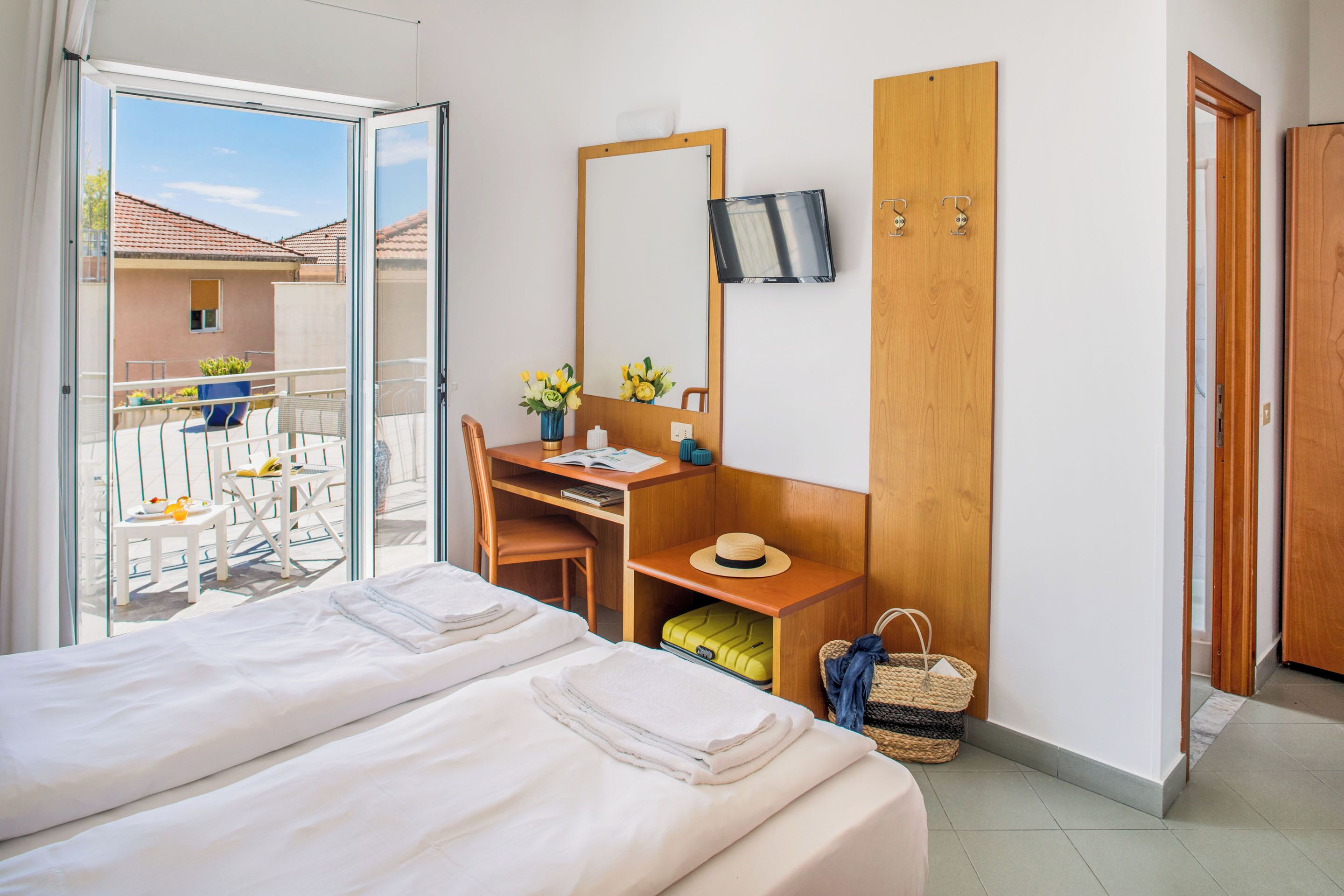 A hotel room with a bed, a desk with a chair, a TV mounted on the wall, and a balcony with outdoor furniture.
