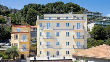Un edificio de varios pisos con balcones, un automóvil estacionado en la parte delantera y una colina bordeada de árboles al fondo.