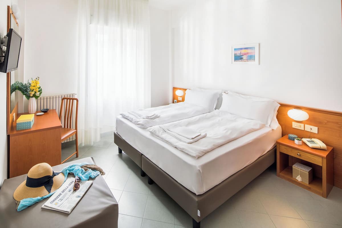 A hotel room with a double bed, bedside tables, a desk with a chair, a TV, and a window with curtains.