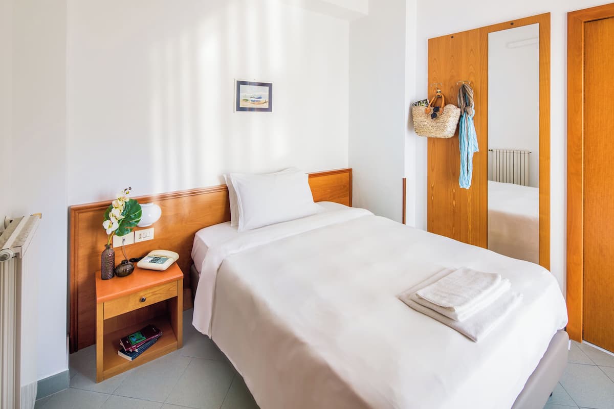A neatly made bed with a white comforter and pillows, a bedside table with a lamp, a phone, and a small plant. There is a wooden wardrobe with a mirror and a hanging basket with a towel.