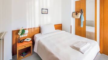 A neatly made bed with a white comforter and pillows, a bedside table with a lamp, a phone, and a small plant. There is a wooden wardrobe with a mirror and a hanging basket with a towel.