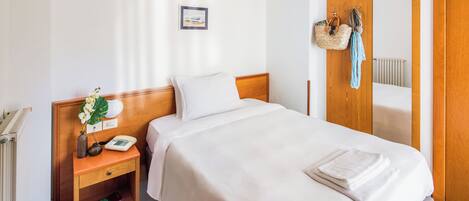 A neatly made bed with a white comforter and pillows, a bedside table with a lamp, a phone, and a small plant. There is a wooden wardrobe with a mirror and a hanging basket with a towel.