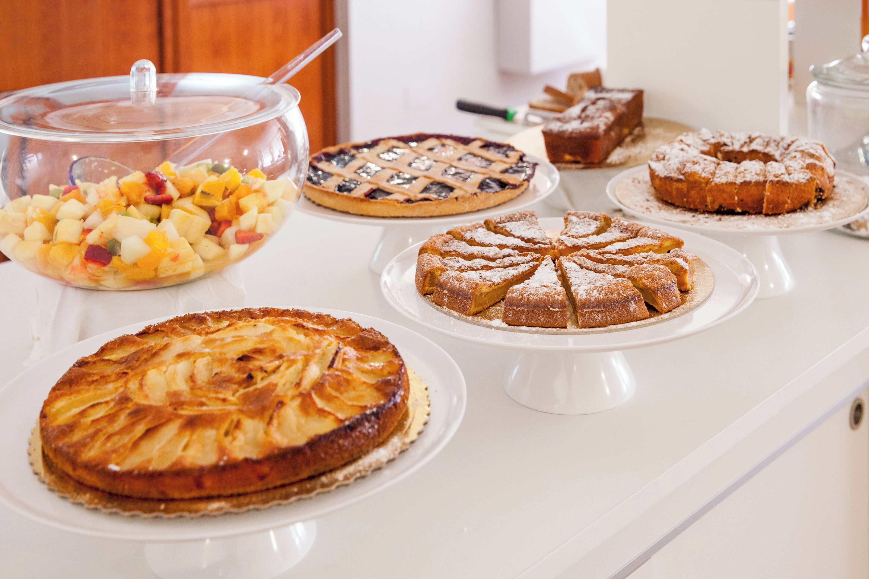 A variety of desserts on a white countertop, including a fruit salad, a pie, a bundt cake, and a cake with a lattice crust.