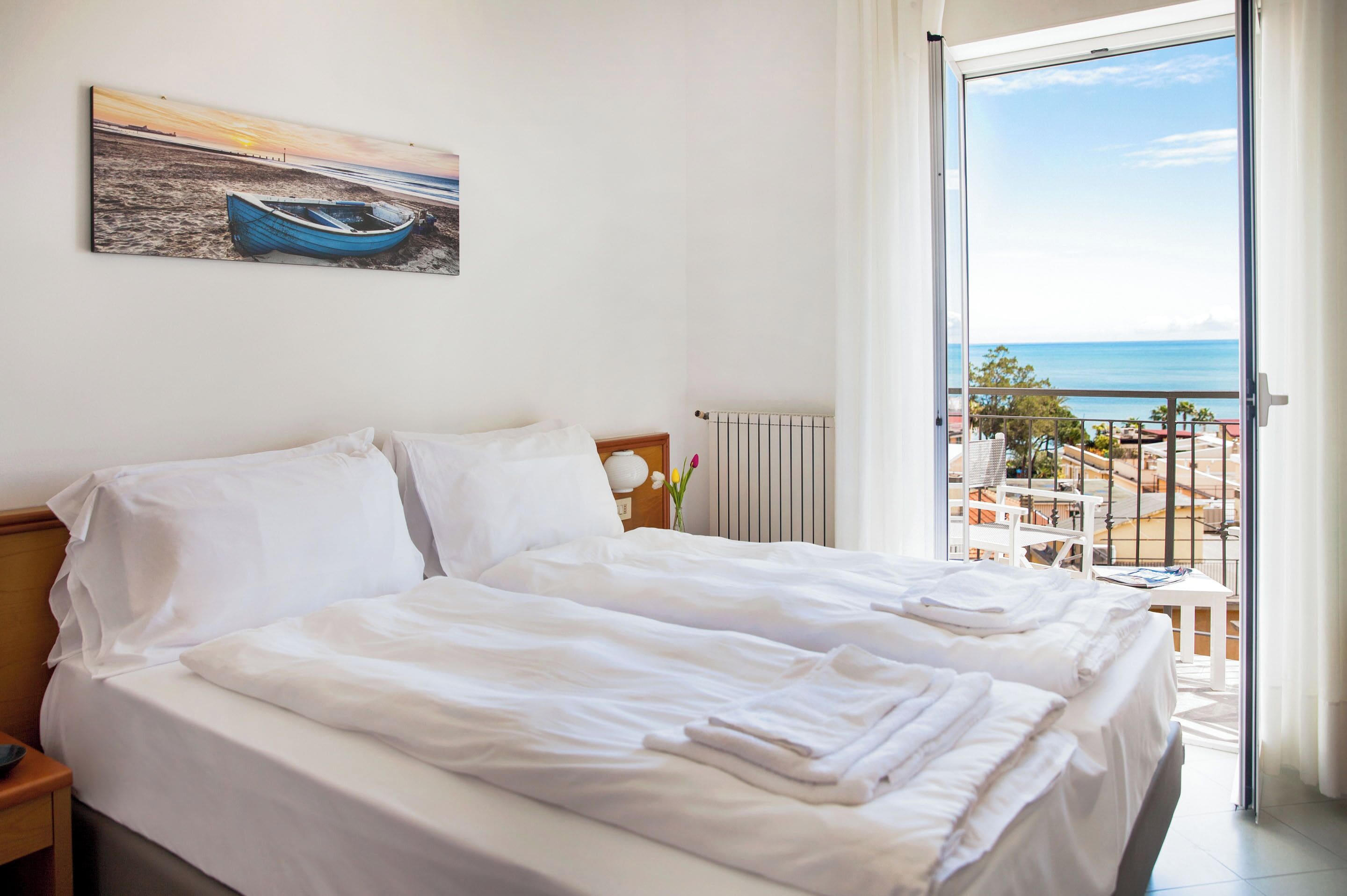 A hotel room with a bed, a nightstand, a window with a view of the sea, and a painting of a boat on the wall.