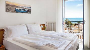 A hotel room with a bed, a nightstand, a window with a view of the sea, and a painting of a boat on the wall.