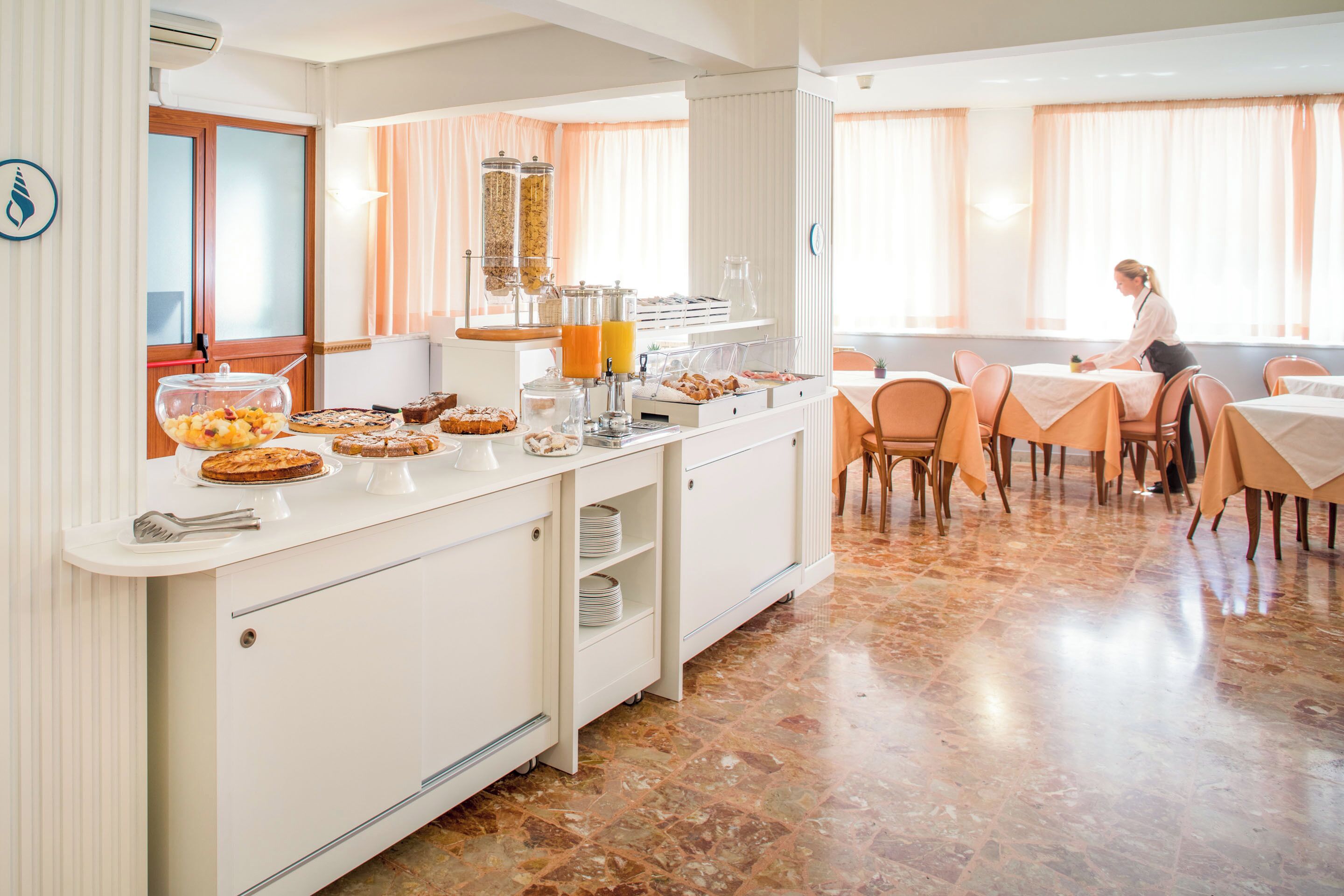 Zona de desayuno tipo buffet con una variedad de pasteles, un dispensador de zumo y una estación de café.