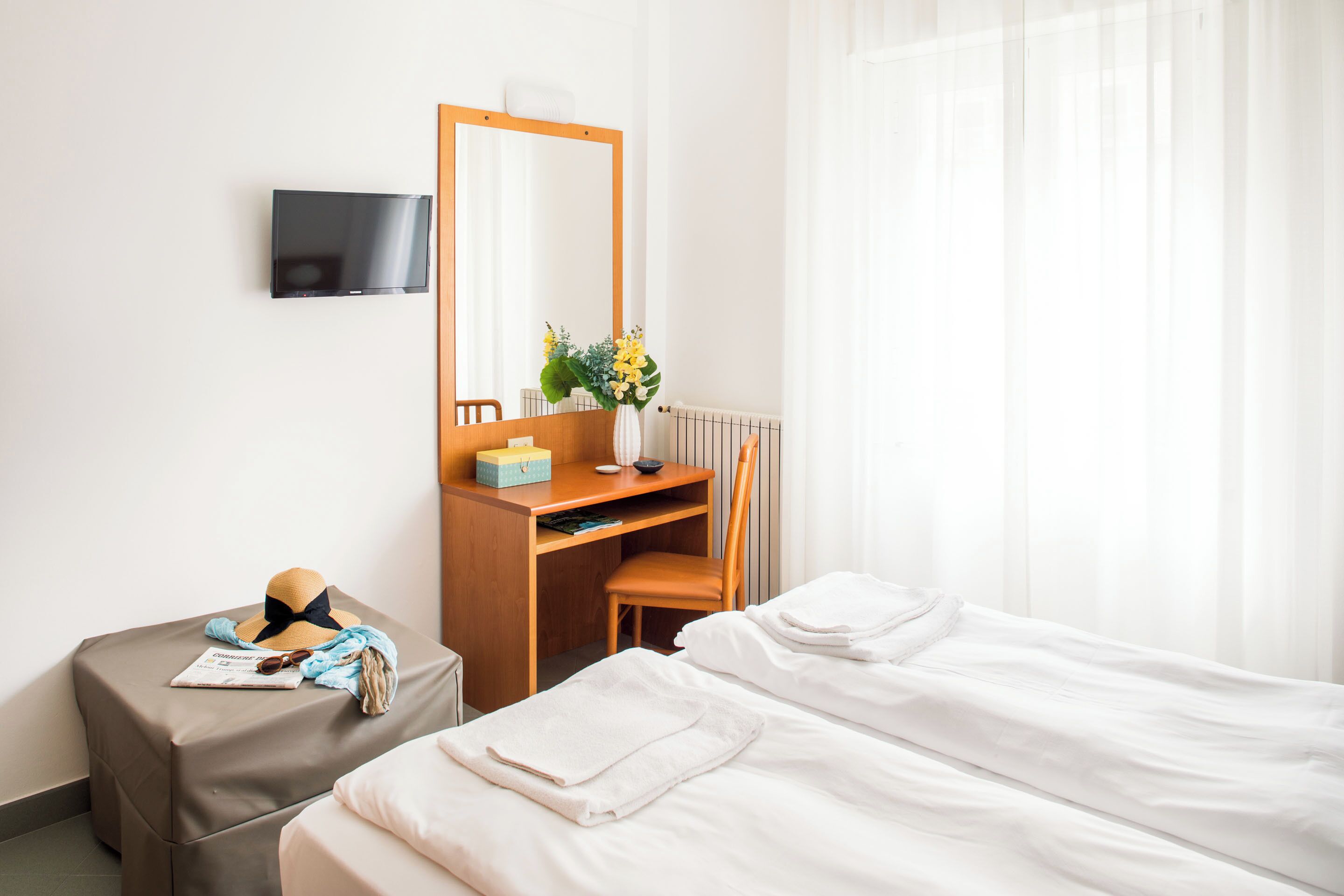 A hotel room with a bed, a desk with a chair, a TV mounted on the wall, and a window with sheer curtains.