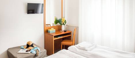 A hotel room with a bed, a desk with a chair, a TV mounted on the wall, and a window with sheer curtains.