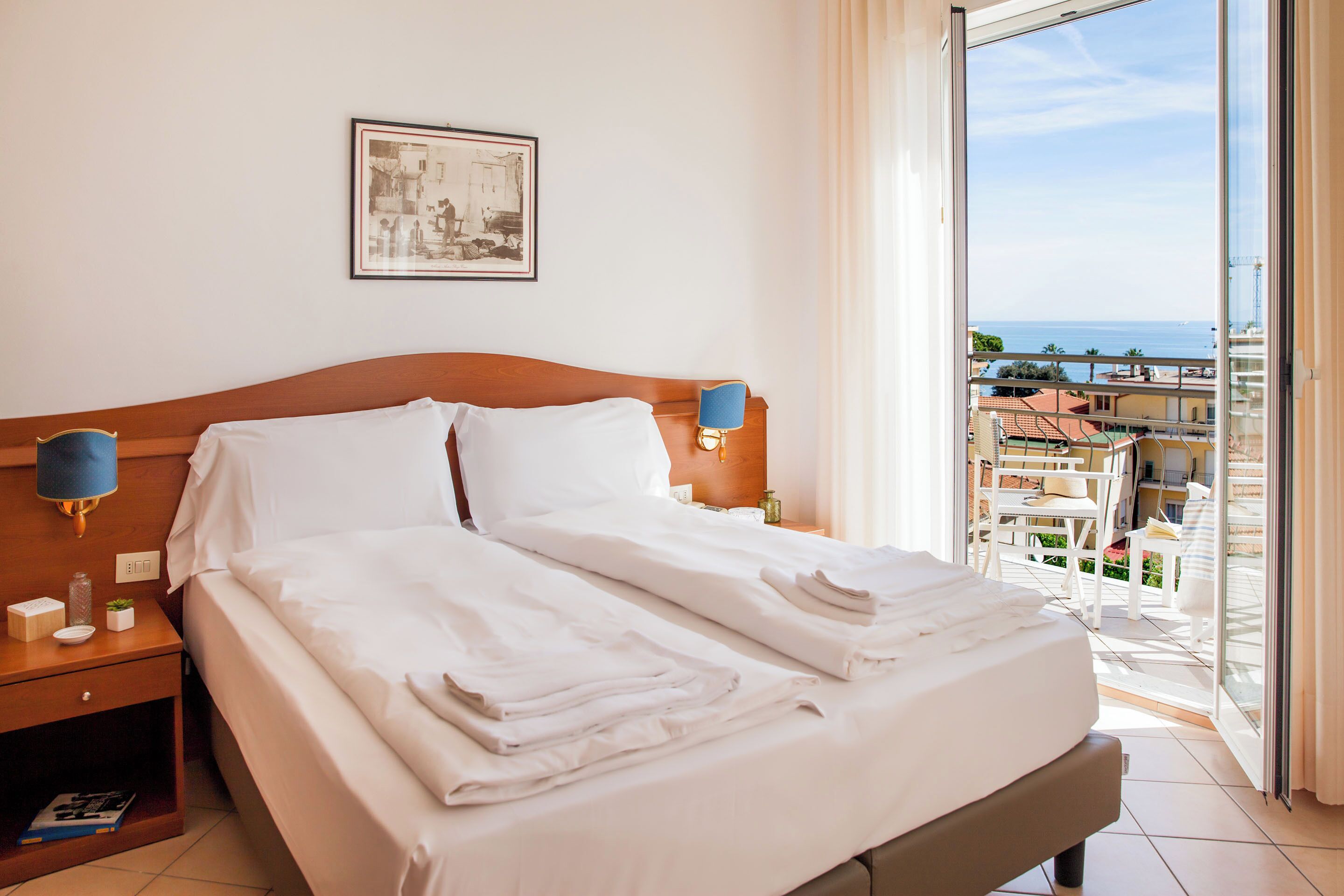A hotel room with a large bed, two bedside lamps, a framed picture on the wall, and a balcony with a view of the sea and buildings.